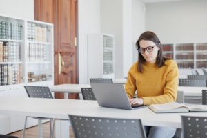 Female graduate student sits at work table in library with her laptop in front of her working on formatting her dissertation