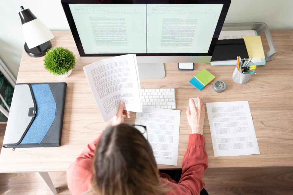 Overhead view of a female graduate student sitting at her desktop computer comparing text on the screen to a paper in her hand as she works on preparing her dissertation for submission.