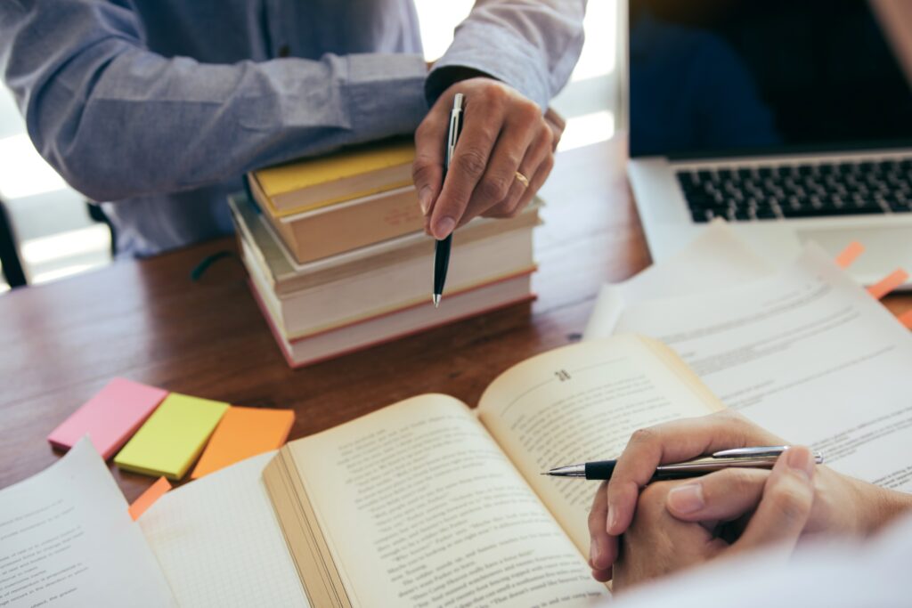 Close up on two men's hands sitting across from each other. One man has his arms resting on a pile of books holding a pen, the other man has a book open on top of his printed dissertation, with his hand resting on it also holding a pen