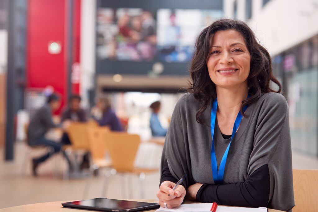A female professor looks at the camera smiling, sitting at a table in the middle of a college building with other students sitting at tables in the background